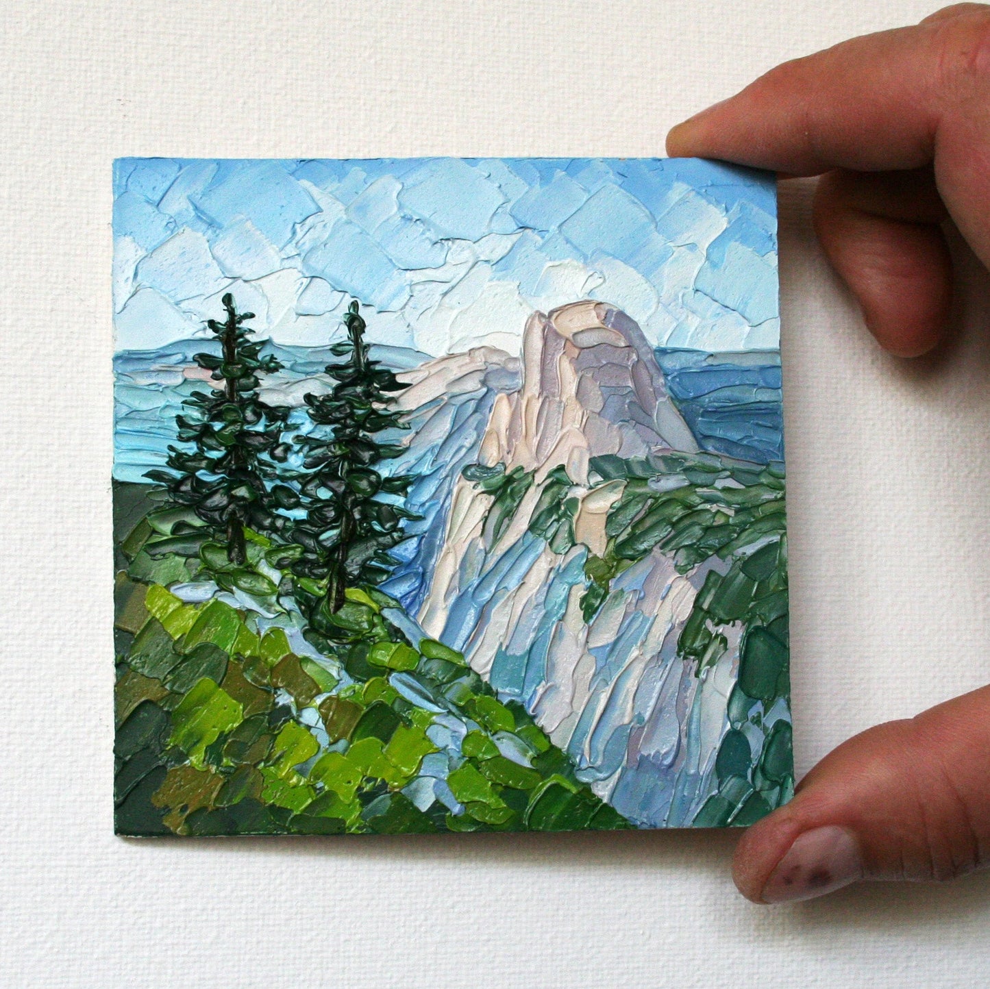 Hand holding a small painting of a mountain landscape with trees.