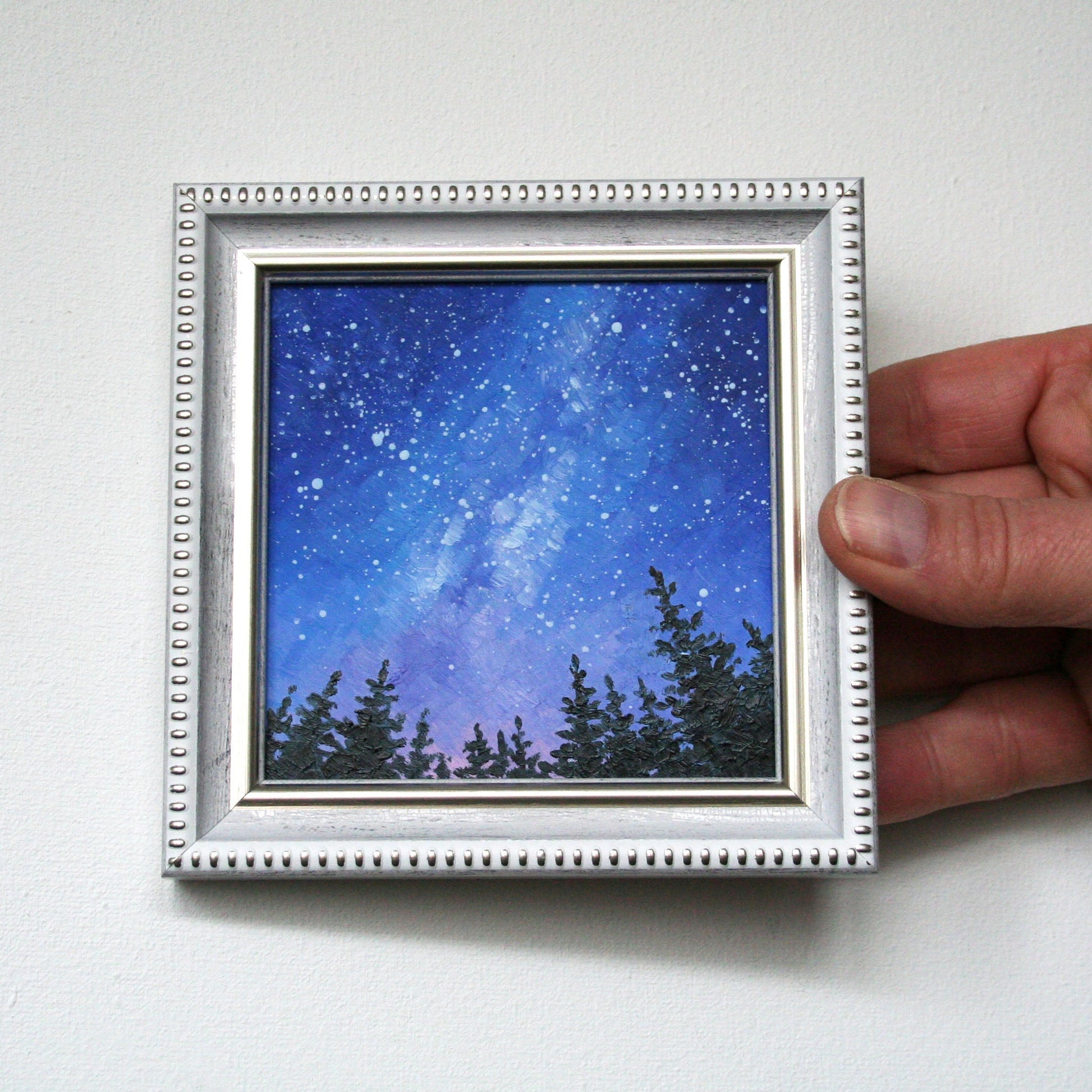 Hand holding a small framed artwork of a night sky with trees.