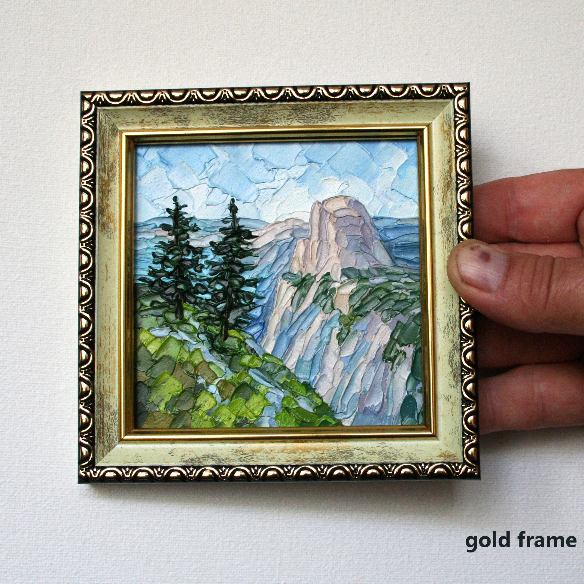 Hand holding a small framed painting of a mountain landscape with a gold frame.