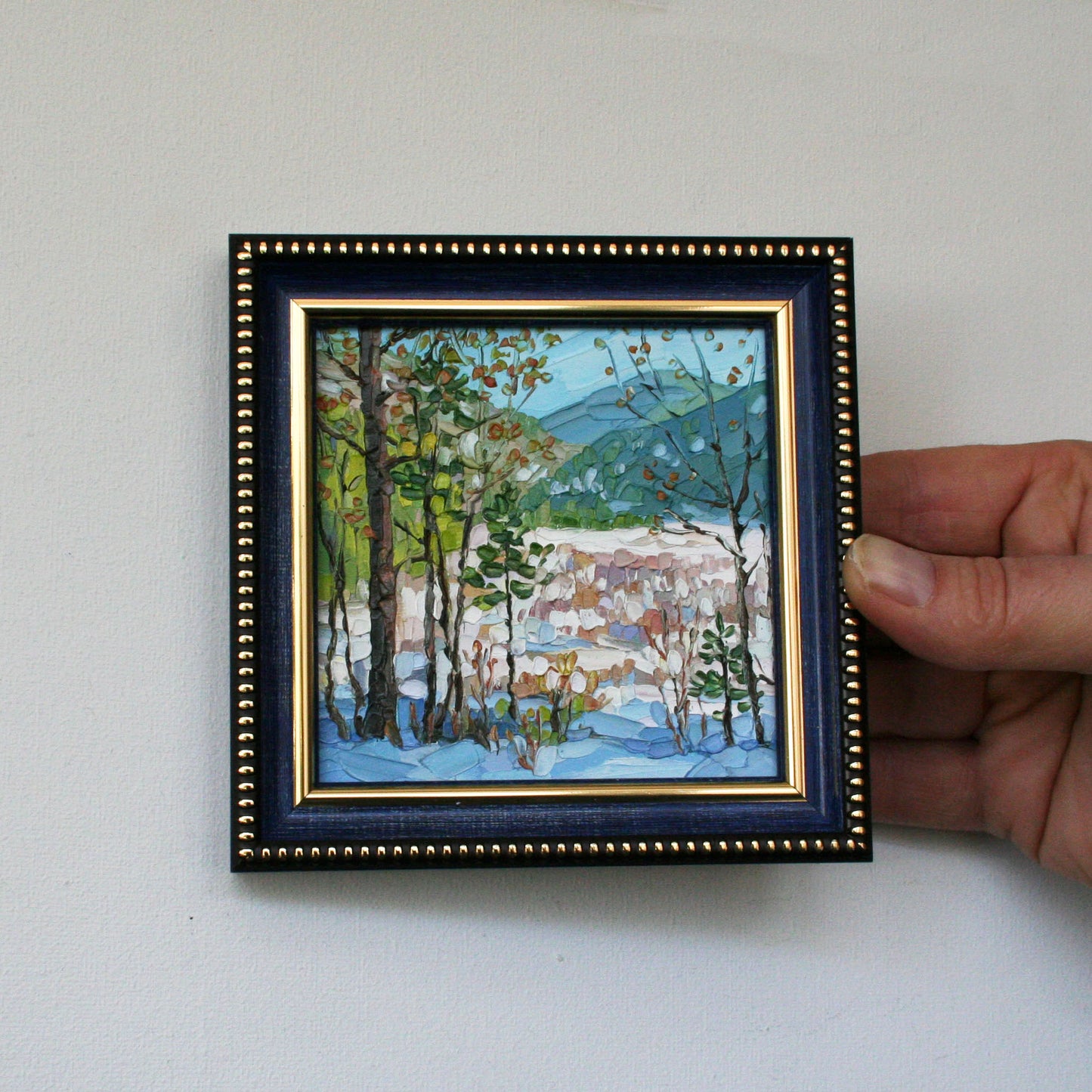 Hand holding a small framed painting of a snowy landscape with trees.