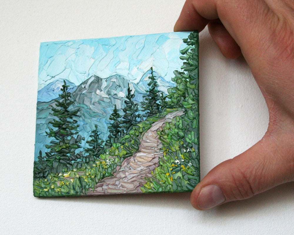 Hand holding a small square painting of a mountain landscape with a path and trees.