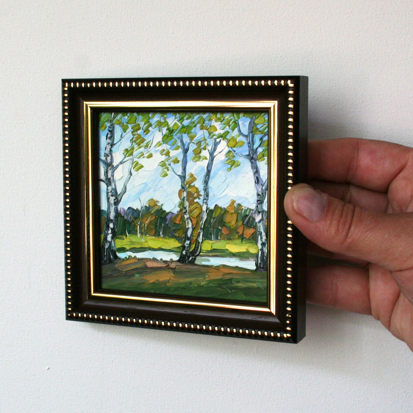 Hand holding a small framed painting of a landscape with trees and a lake.