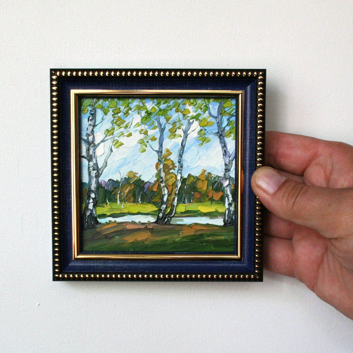 Hand holding a small framed painting of a landscape with trees.