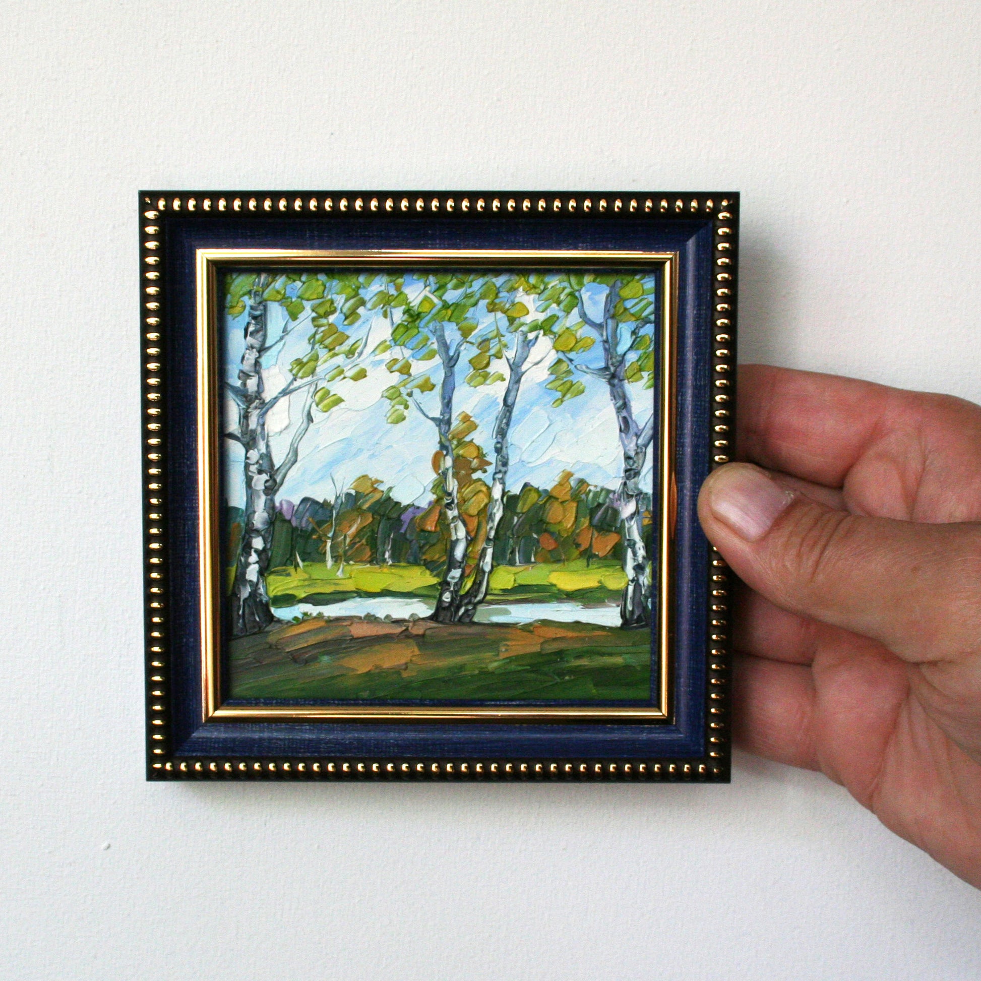 Hand holding a small framed painting of a landscape with trees.