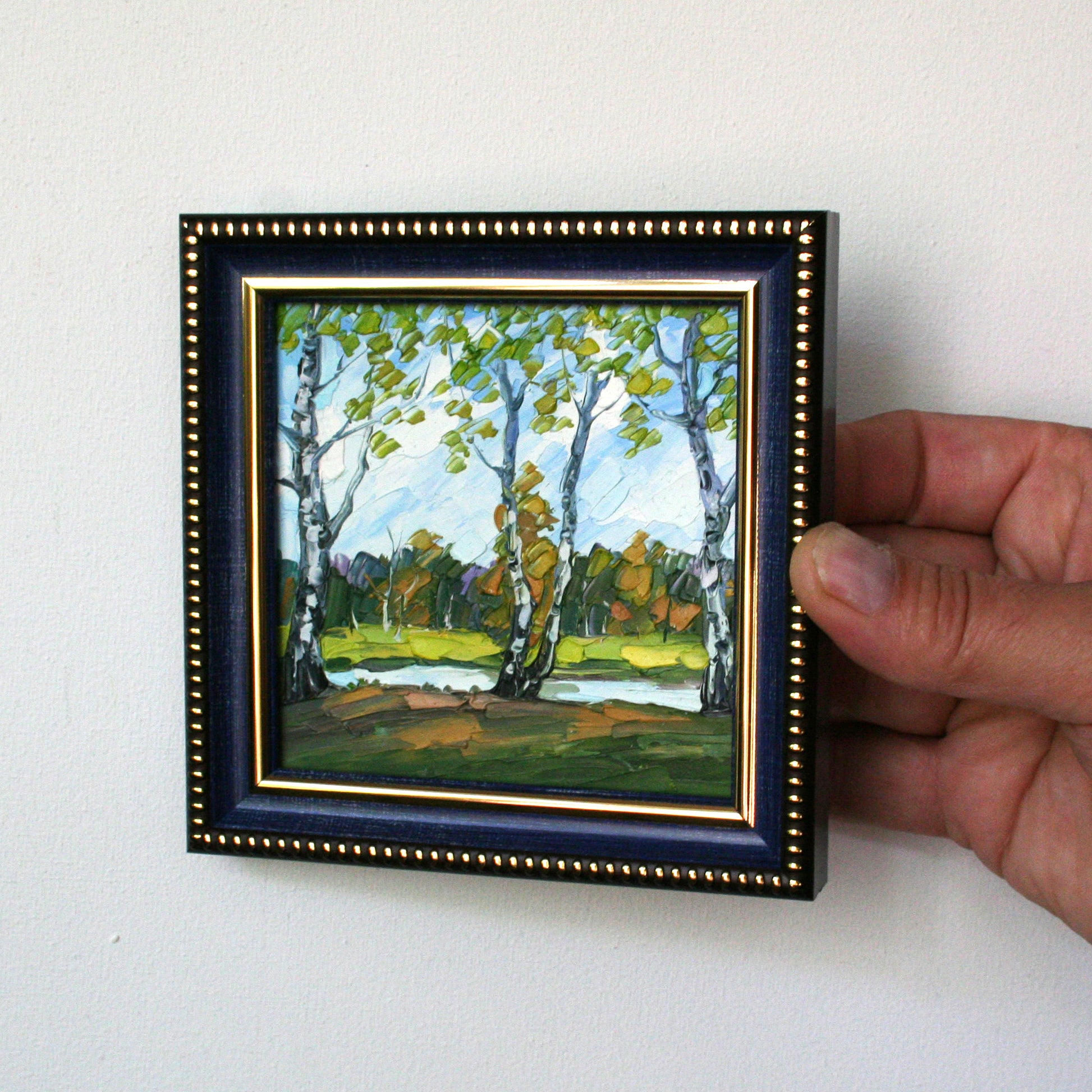 Hand holding a small framed painting of a landscape with trees.