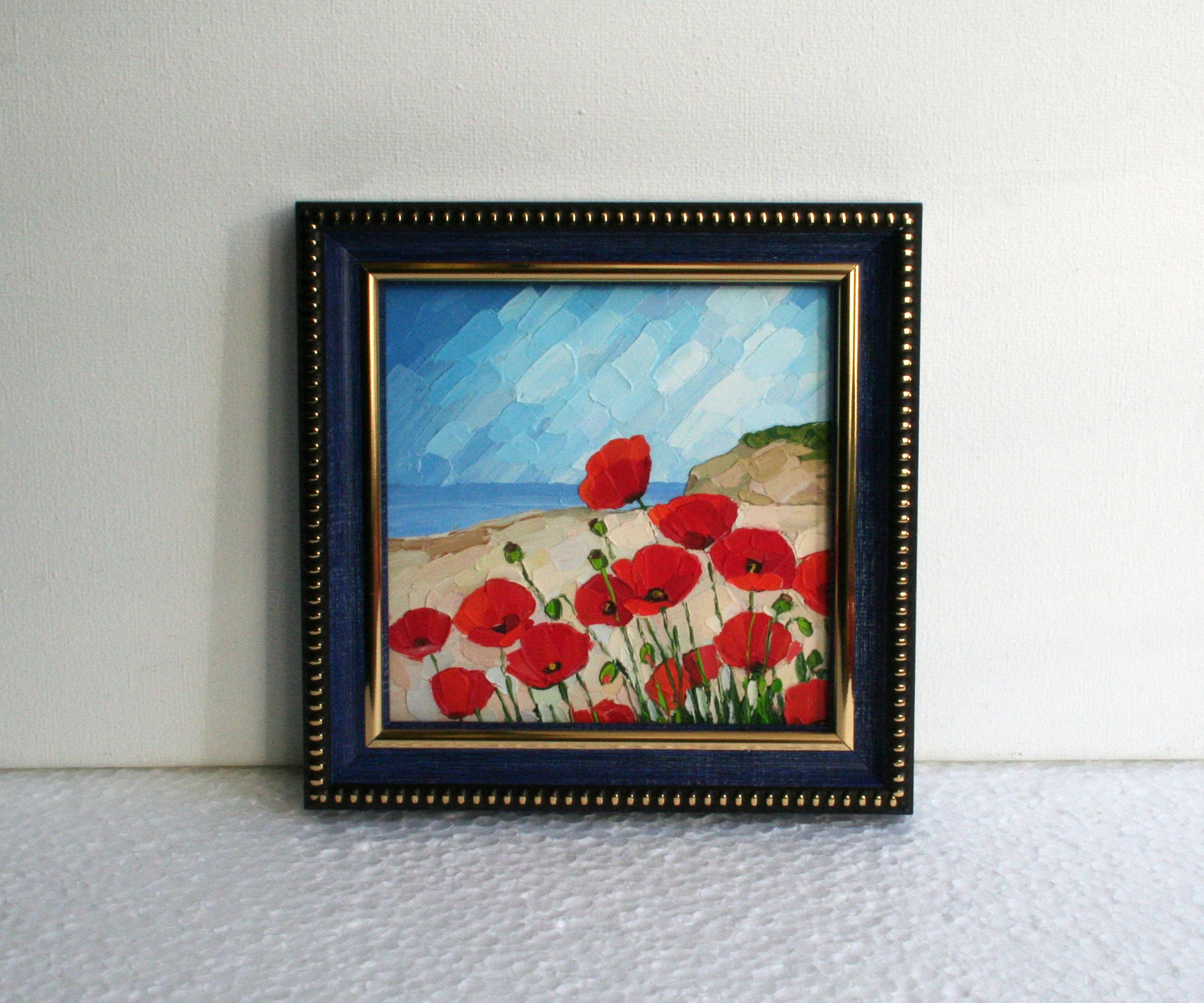 Framed painting of red flowers on a beach with a blue sky.