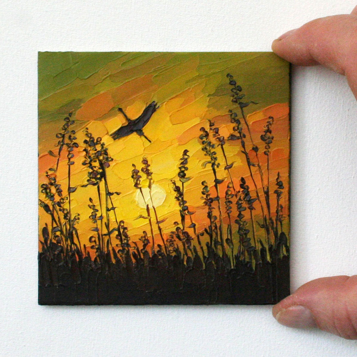Small painting of a sunset scene with silhouetted plants and a bird, held by a hand.