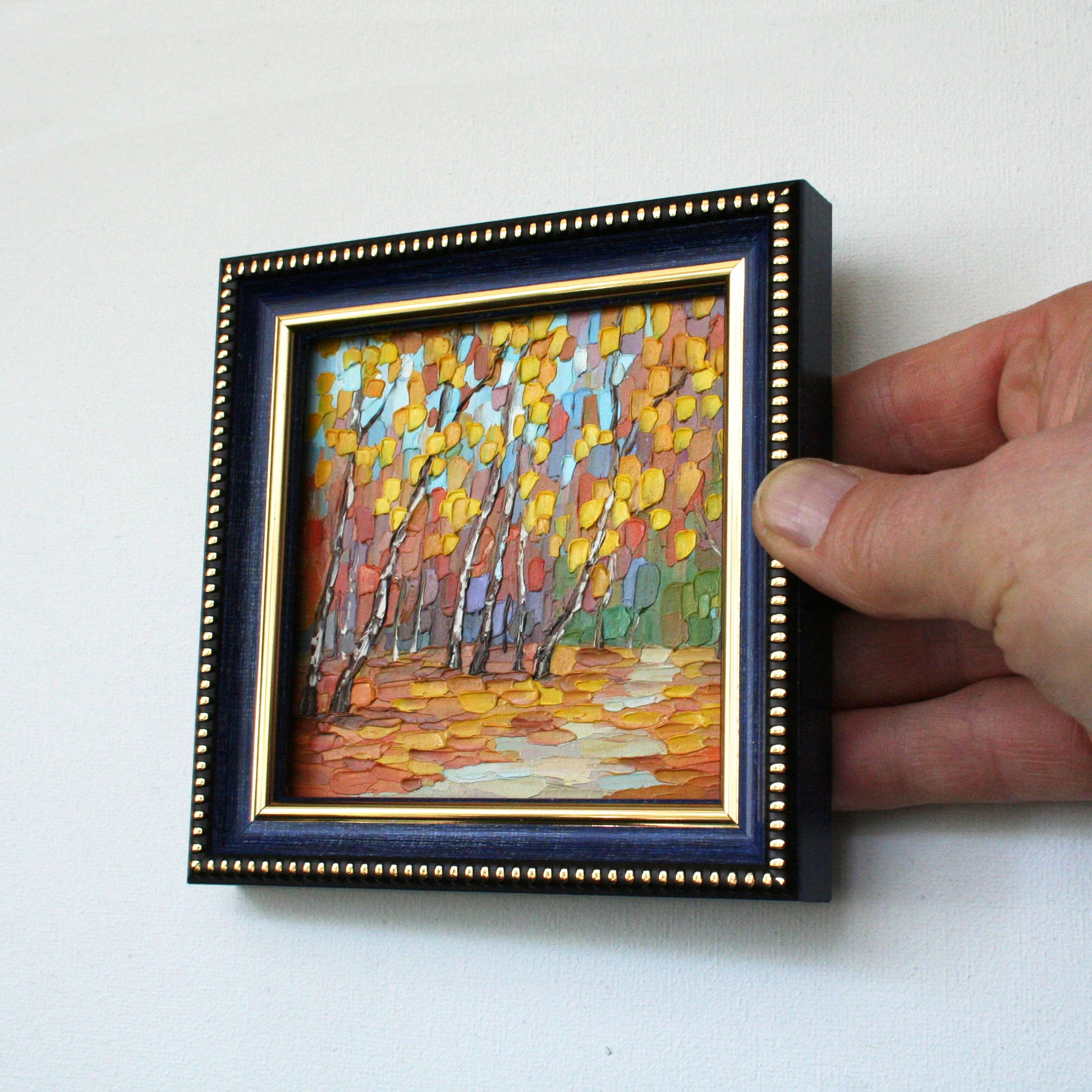 Hand holding a small framed painting of a landscape with trees.