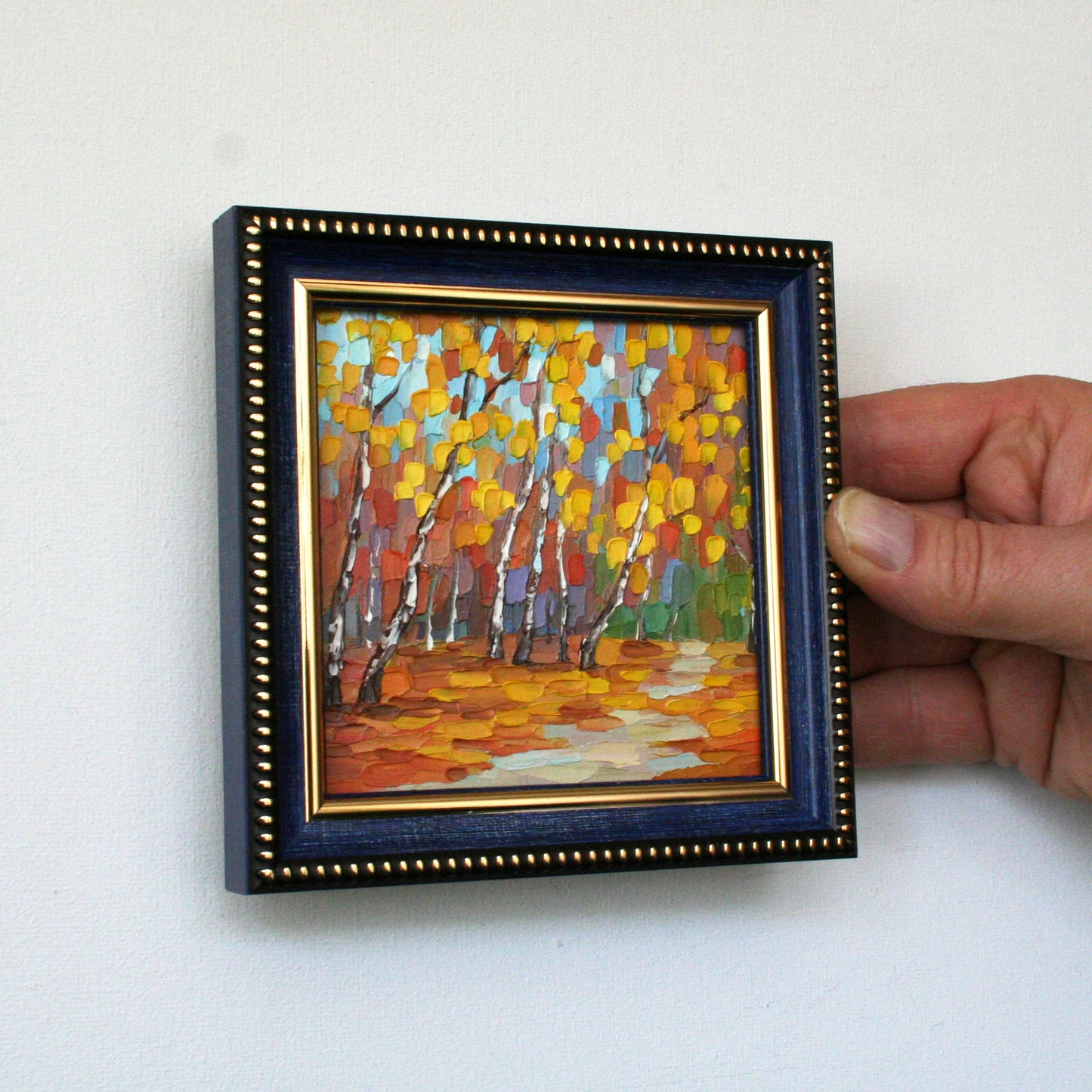 Hand holding a small framed painting of a forest scene with autumn colors.